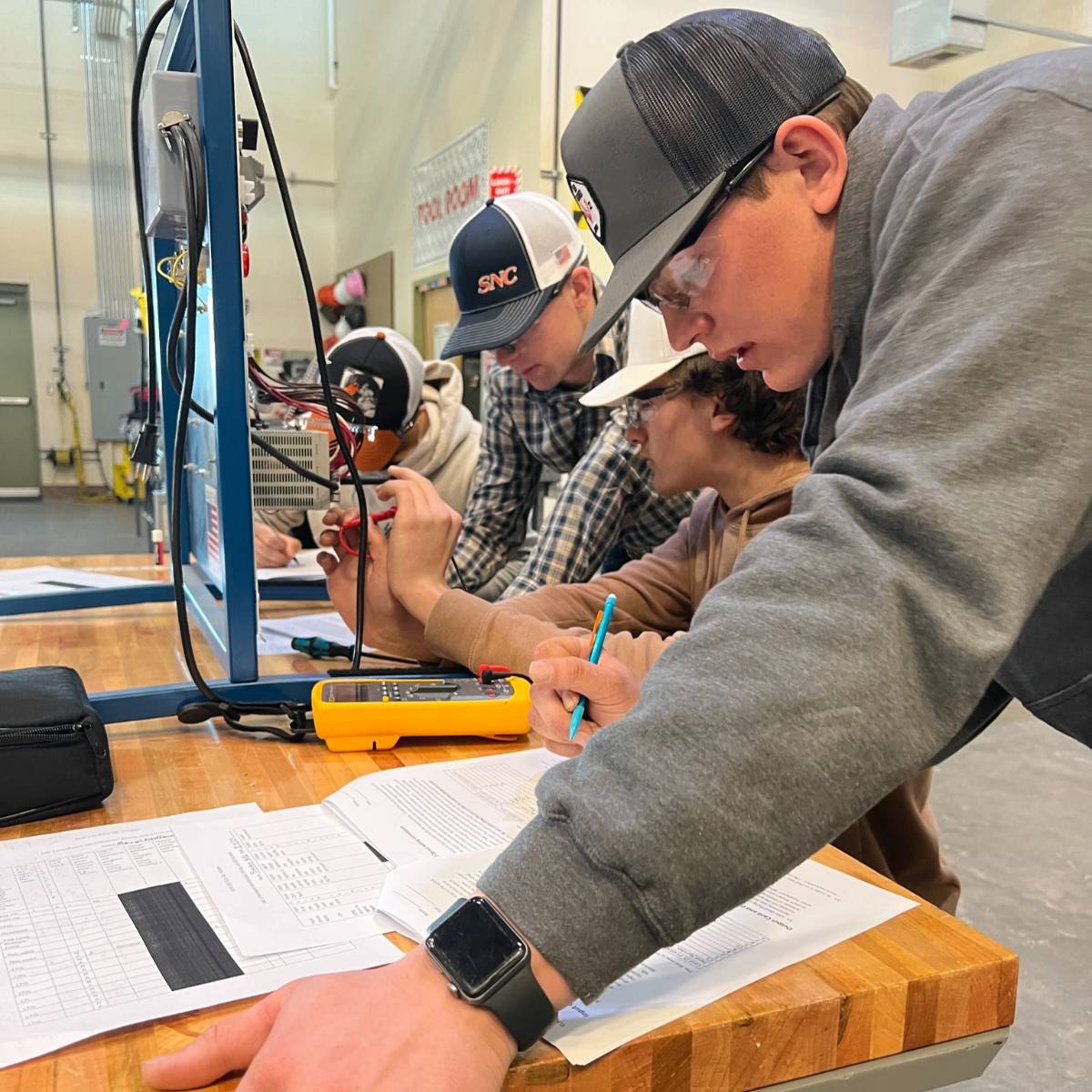 CTE students working together at a table with an electrical panel, meter, and papers for calculations.